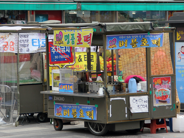 A bustling Myeongdong street scene with various colorful Korean street food stalls.